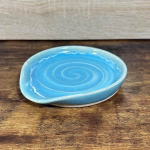 May include: A handmade ceramic spoon rest with a light blue glaze. The spoon rest features a spiral design in the center and a wavy edge. The base is white. The spoon rest is on a wooden surface.