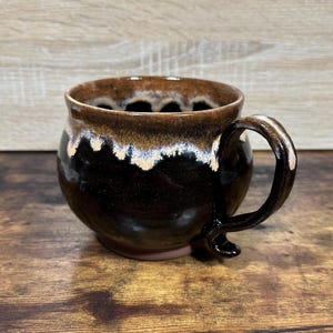 May include: A handmade ceramic mug with a dark brown glaze, accented with lighter brown and white details around the rim. The mug has a rounded shape and a curved handle, perfect for enjoying coffee or tea.