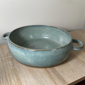 May include: A light blue ceramic serving bowl with two handles. The bowl has a speckled glaze and a brown rim.