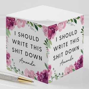 May include: White notepad cube with floral accents and the text "I SHOULD WRITE THIS SHIT DOWN Amanda". The design includes pink and purple flowers and green leaves. A pen with a gold tip is in the foreground.