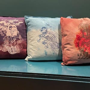 May include: Three decorative throw pillows with different designs. The pillow on the left has a purple and white design with a creature in the center. The middle pillow has a blue and white design with a creature in the center. The pillow on the right has a pink and red design with a creature in the center.