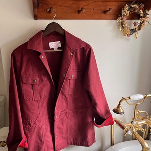May include: A burgundy jacket with four front pockets and a collared neckline hangs on a wooden hanger. The jacket's cuffs are lined with a red and black plaid pattern. The jacket is displayed in a bathroom setting.