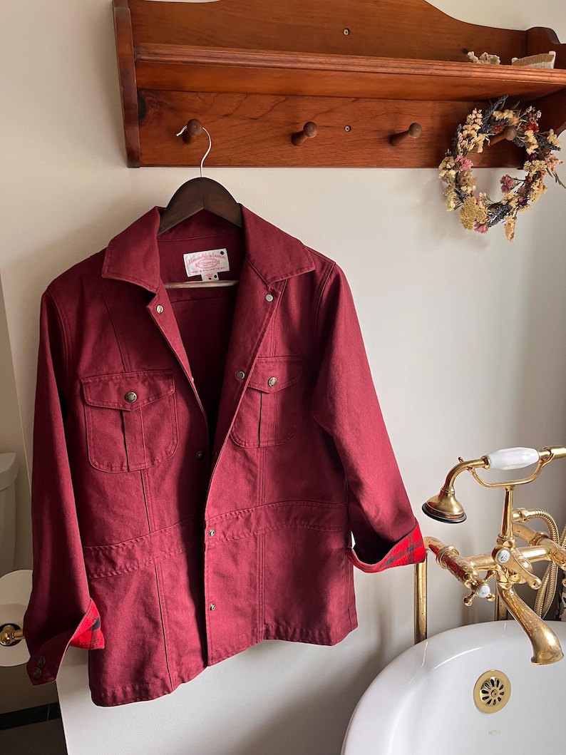 May include: A burgundy jacket with four front pockets and a collared neckline hangs on a wooden hanger. The jacket's cuffs are lined with a red and black plaid pattern. The jacket is displayed in a bathroom setting.