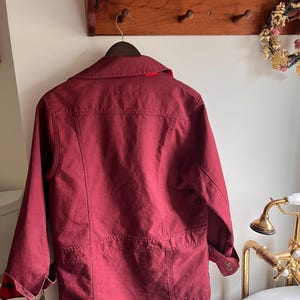 May include: A burgundy jacket with a collared neckline and long sleeves hangs on a wooden hanger. The jacket features a red lining at the collar and plaid detailing at the cuffs. The jacket is hanging on a wooden rack with pegs.
