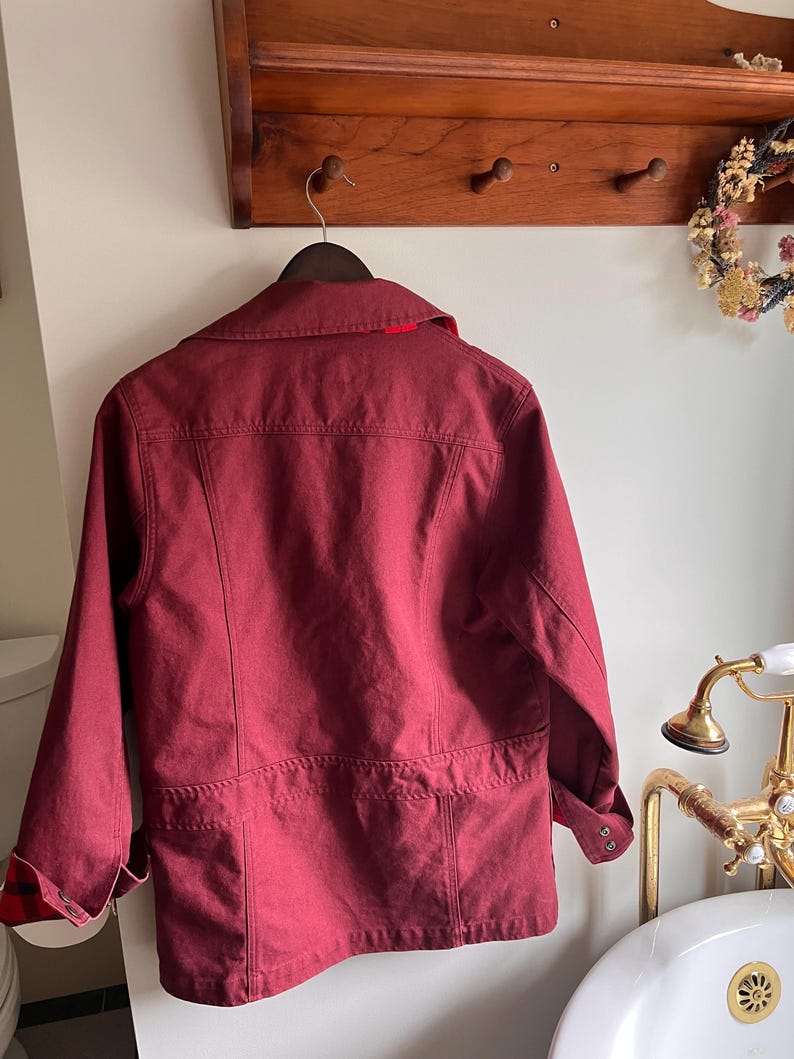 May include: A burgundy jacket with a collared neckline and long sleeves hangs on a wooden hanger. The jacket features a red lining at the collar and plaid detailing at the cuffs. The jacket is hanging on a wooden rack with pegs.