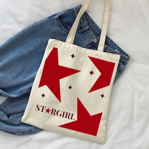 May include: Cream-colored tote bag with red star and diamond designs, and the text "STARGIRL". The bag has a rectangular shape with a handle and is placed on top of blue jeans.