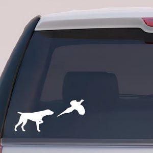 Puede incluir: Calcomanía de vinilo blanco con un perro de caza y un faisán en vuelo. El perro está en posición de muestra, y el faisán está en pleno vuelo. Diseñado para ser aplicado en la ventana de un vehículo.