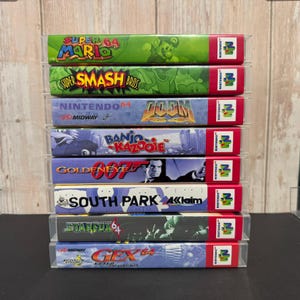 May include: A vertical stack of eight Nintendo 64 video game boxes in clear protective cases. Titles include Super Mario 64, Super Smash Bros, Doom 64, Banjo-Kazooie, GoldenEye 007, South Park, Starfox 64, and Gex 64: Enter the Gecko, all featuring the N64 logo.