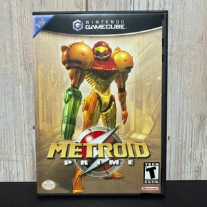 May include: A Nintendo GameCube video game case for Metroid Prime featuring a central illustration of the character Samus Aran in orange and yellow armor with a green arm cannon. The title Metroid Prime is printed in gold with a red circular emblem.
