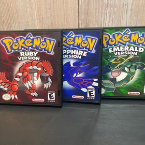 May include: Three Game Boy Advance video game cartridges for Pokemon Ruby, Sapphire, and Emerald versions. The cartridges are black with colorful artwork featuring the Pokemon characters.
