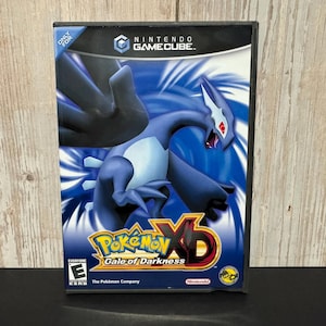 May include: A Nintendo GameCube video game case for Pokémon XD: Gale of Darkness. The cover features a dark blue Lugia creature with red eyes against a blue and white swirling background. The title is printed in yellow and red text at the bottom.