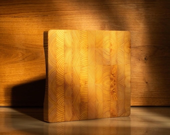May include: A square, end-grain wooden chopping board with a light-coloured, natural wood grain pattern. The board is lit by a warm light, casting a shadow on the background. The board is on a marble surface.