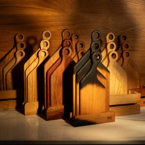 Könnte beinhalten: Eine Sammlung von Holzschneidebrettern in verschiedenen Braun- und Naturholztönen. Einige Bretter haben eine dunkle, verkohlte Oberfläche. Jedes Brett hat einen Griff mit einer kreisförmigen Öffnung. Die Bretter stehen auf einer weißen Oberfläche.