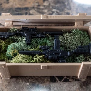May include: Two black toy guns are displayed in a wooden crate lined with green moss. The crate is open and the guns are visible.