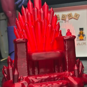 May include: A red, 3D-printed throne with a crystal-like backrest. The throne features a seat, armrests, and a base with additional crystal formations. The overall design is a fantasy-themed decorative item.
