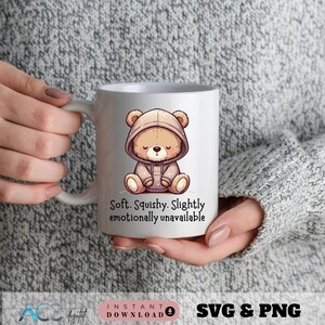 May include: White ceramic mug featuring a cartoon bear in a hoodie. The bear is light brown with a closed-eye expression. Text on the mug reads: "Soft. Squishy. Slightly emotionally unavailable."
