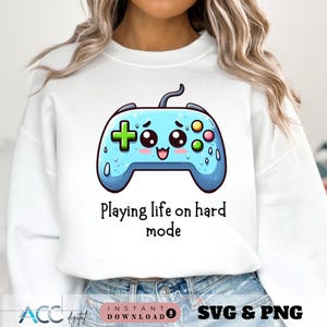 May include: White sweatshirt featuring a cartoon-style light blue video game controller with a smiling face. The text "Playing life on hard mode" is below the controller. The design includes a plus sign, colorful buttons, and water droplet details.