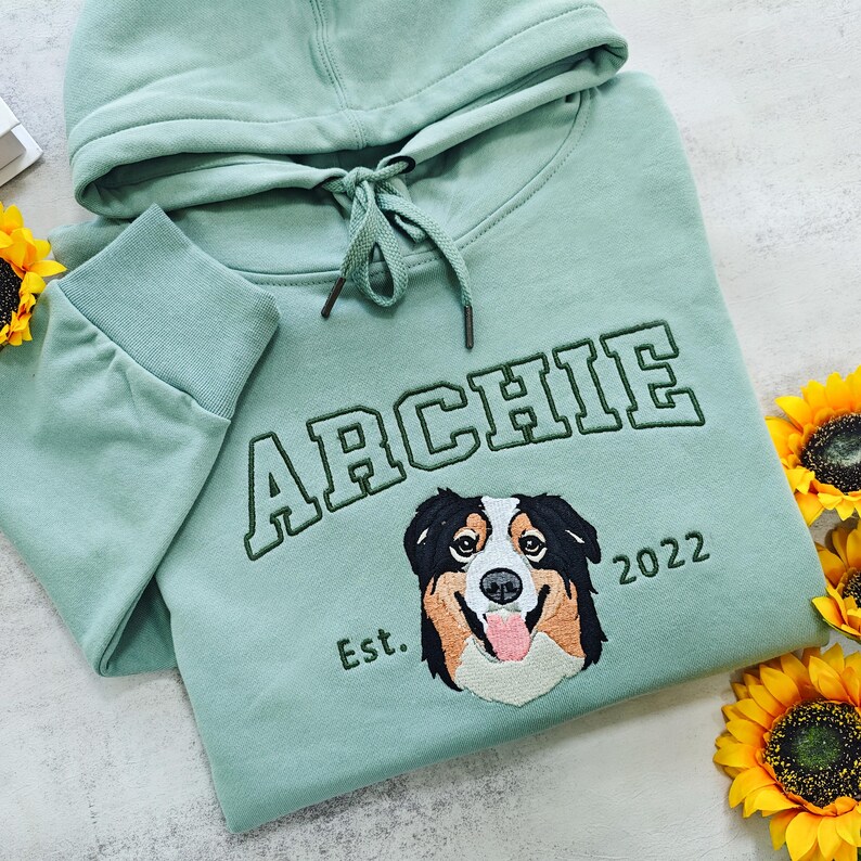 May include: A light green hooded sweatshirt with the word "ARCHIE" embroidered in black and green, along with a dog's portrait and the text "Est. 2022".