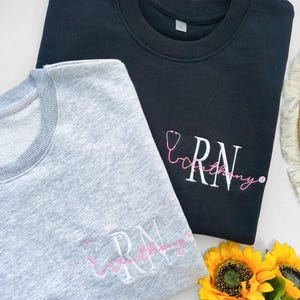 May include: Two sweatshirts, one grey and one black, with the text "RN" embroidered on each. The text is accompanied by a pink embroidered stethoscope.