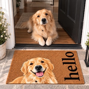 Puede incluir: Un felpudo rectangular de fibra de coco marrón con una ilustración impresa de un perro golden retriever con la boca abierta y la lengua fuera. La palabra hello está impresa en letras minúsculas negras en el lado derecho de la alfombra.