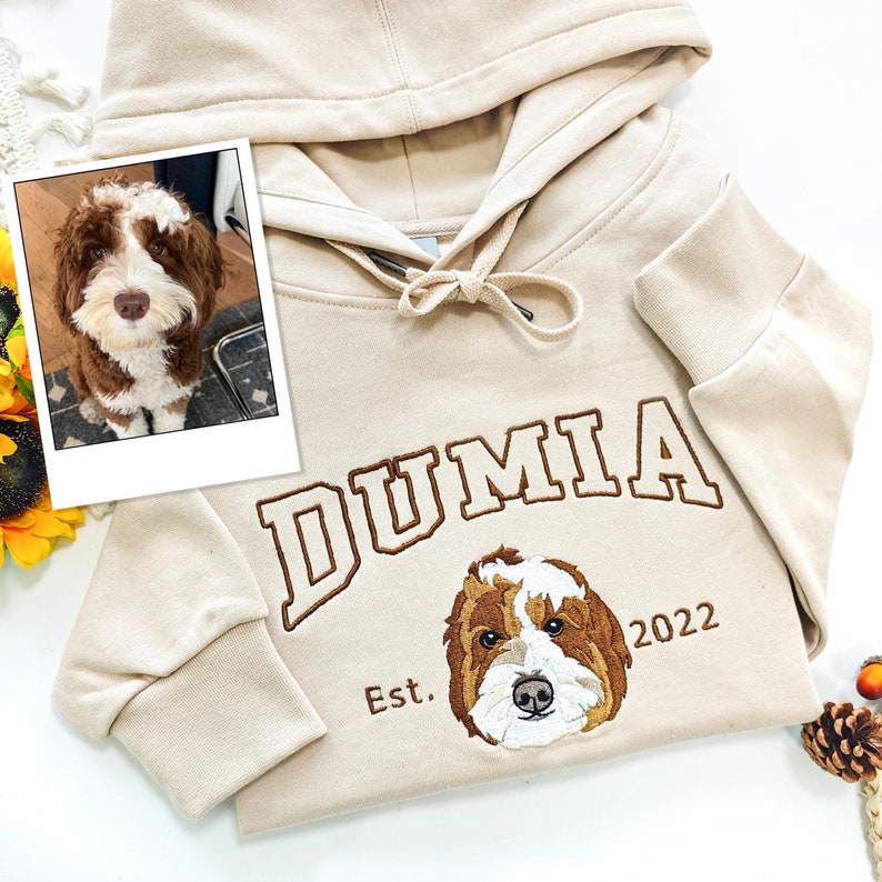 May include: A beige hoodie with a brown embroidered dog and the text "DUMIA Est. 2022".