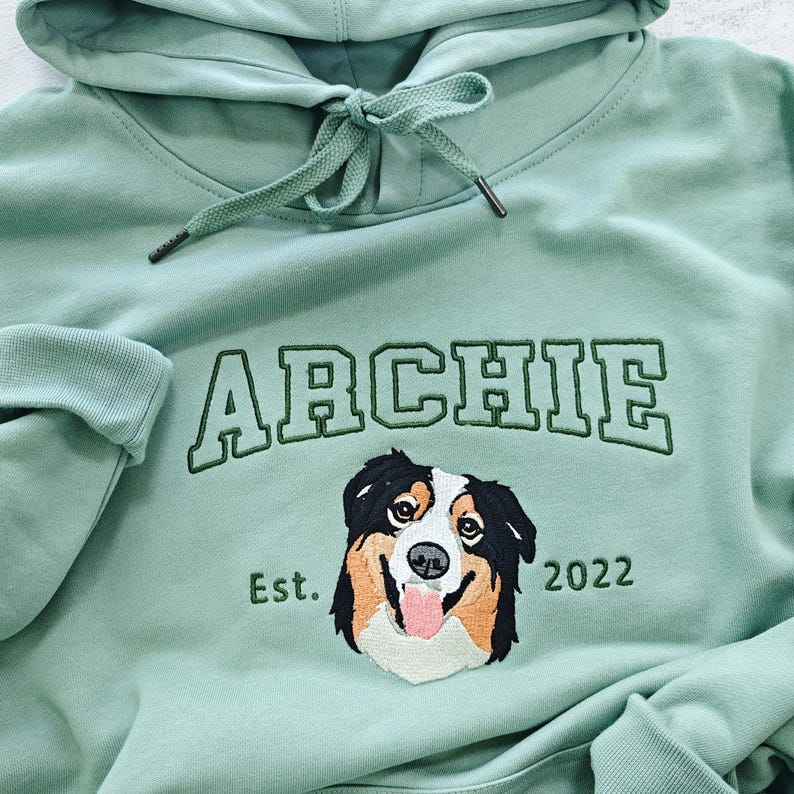 May include: A light green hooded sweatshirt with the name "ARCHIE" embroidered in white with a green outline.  Below the name is a black and white dog with brown markings, and the text "Est. 2022".