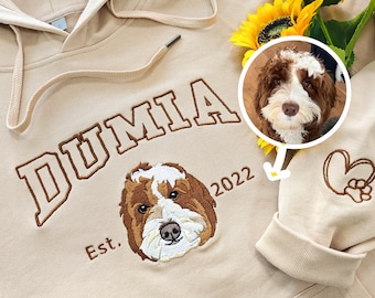 May include: A beige hoodie with the word "DUMIA" embroidered in brown on the front. The hoodie also has a brown embroidered dog portrait and the text "Est. 2022".