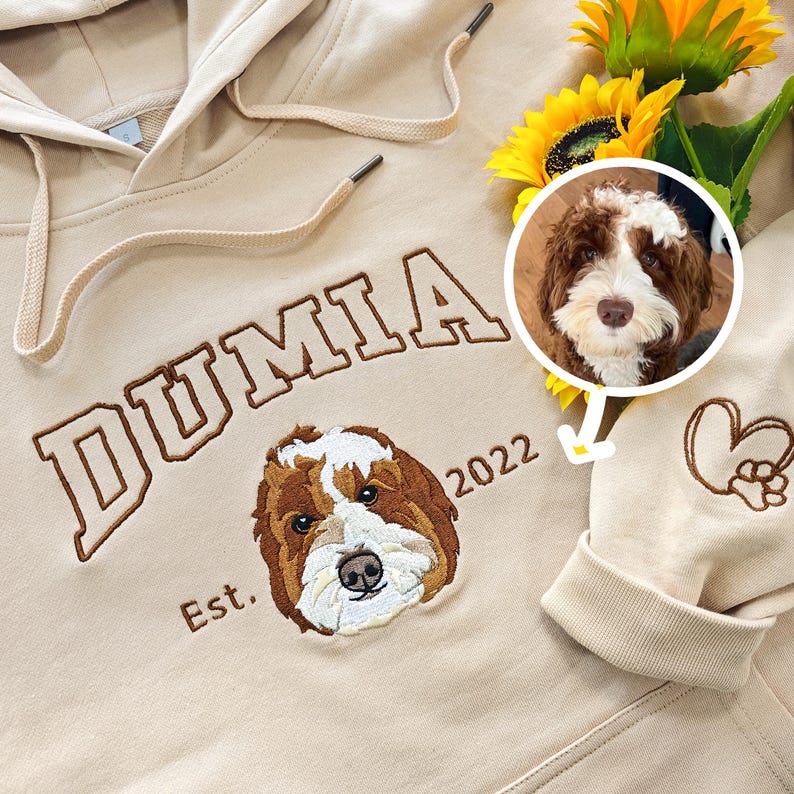 May include: A beige hoodie with the word "DUMIA" embroidered in brown on the front. The hoodie also has a brown embroidered dog portrait and the text "Est. 2022".