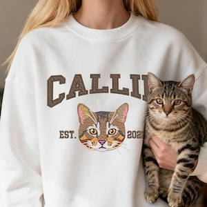 May include: Cream-coloured sweatshirt featuring the name "CALLI" in brown lettering above a cat face design. The graphic includes the text "EST. 2021". A tabby cat is held in the image.