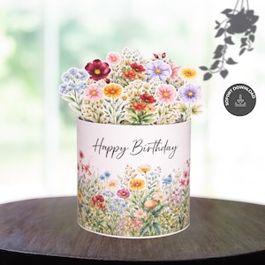 Könnte beinhalten: Eine Pop-up-Geburtstagskarte mit einem floralen Arrangement. Die zylindrische Karte hat eine weiße Basis mit bunten Blumen und dem Schriftzug "Happy Birthday". Die Oberseite zeigt einen Strauß Papierblumen in verschiedenen Farben.