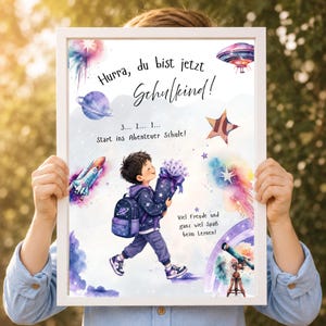 Puede incluir: Una impresión enmarcada con una ilustración en acuarela de un niño con mochila y un cono de regalo, rodeado de elementos espaciales como planetas, un cohete y un telescopio. El texto dice: Hurra, du bist jetzt Schulkind! 3... 2... 1... Start ins Abenteuer Schule! Viel Freude und ganz viel Spaß beim Lernen!