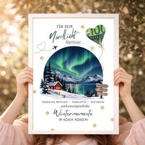 Op de afbeelding: Een ingelijste print met een cirkelvormige illustratie van een besneeuwd landschap met een rode hut, bergen en groen noorderlicht. Het bevat Duitse tekst, een hartvormige ballon met een briefje van 100 euro en gouden sneeuwvlokken.