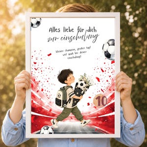 Puede incluir: Una impresión enmarcada con una ilustración de dibujos animados de un niño con mochila que sostiene un ramo de balones de fútbol. El texto dice: Alles liebe für dich zur einschulung, kleiner champion, großer tag! viel spaß bei deiner einschulung! Se ven detalles de confeti rojo y el número 10.