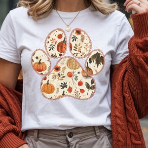 May include: White t-shirt featuring a paw print design filled with fall-themed illustrations. The paw print includes pumpkins, leaves, and floral elements in shades of orange, brown, and beige. The design is outlined with a decorative border.