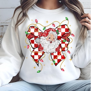 May include: White sweatshirt featuring a festive Santa Claus design. The graphic includes a red and white checkered bow, Santa's face, and string lights. The design is embellished with glitter and gold accents, perfect for the holiday season.