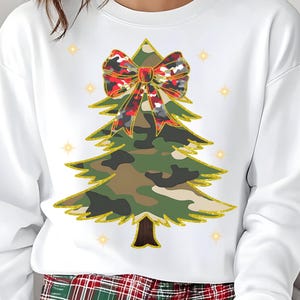 Può includere: Felpa bianca con un design a forma di albero di Natale. L'albero è verde mimetico con finiture glitterate dorate e un fiocco mimetico rosso, nero e bianco. Sono inclusi anche dettagli a stella dorata.