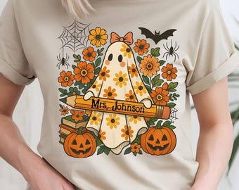 May include: Beige t-shirt with a Halloween design. The graphic shows a ghost holding a pencil with the text "Mrs. Johnson", surrounded by orange flowers, pumpkins, spiders, a bat, and spiderwebs. The ghost is decorated with yellow flowers.