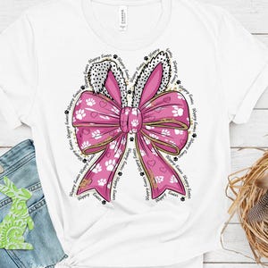 May include: A white t-shirt with a pink bow with white paw prints and gold glitter. The bow has bunny ears and the text "Happy Easter" is repeated around the bow.