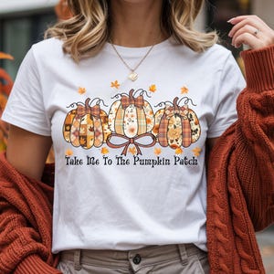 以下が含まれることがあります： チェック柄と花柄の3つの装飾的なカボチャが特徴の白いTシャツ。それぞれにリボンが付いています。「Take Me To The Pumpkin Patch」のテキストが下に印刷されています。秋をテーマにしたアパレル。