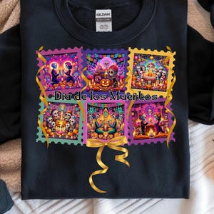May include: Black sweatshirt featuring a colorful Día de los Muertos design. The design includes six postage stamp-style images with sugar skulls, candles, and festive decorations. Gold ribbon accents the design, and the text "Día de los Muertos" is displayed.