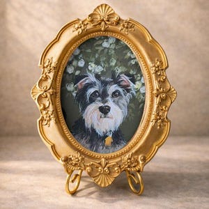 May include: An oval gold-colored ornate picture frame featuring a detailed painting of a black and white terrier-type dog with a yellow tag. The frame has intricate floral carvings and rests on a matching gold-colored metal stand with two curved feet.