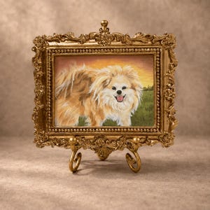 May include: A rectangular painting of a fluffy, light brown and white dog with a happy expression, set within an ornate, metallic gold-colored frame. The artwork is displayed on a matching gold-colored metal stand with curved, decorative feet.