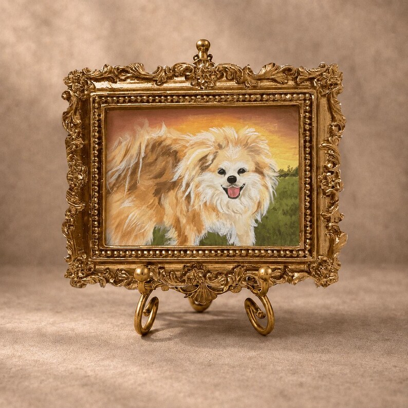 May include: A rectangular painting of a fluffy, light brown and white dog with a happy expression, set within an ornate, metallic gold-colored frame. The artwork is displayed on a matching gold-colored metal stand with curved, decorative feet.