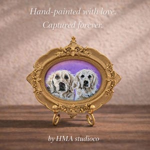 May include: An oval gold-toned ornate frame containing a painting of two light-colored dogs against a purple background. The frame rests on a small metal stand. Text above reads: Hand-painted with love. Captured forever. Text below reads: by HMA studioco.