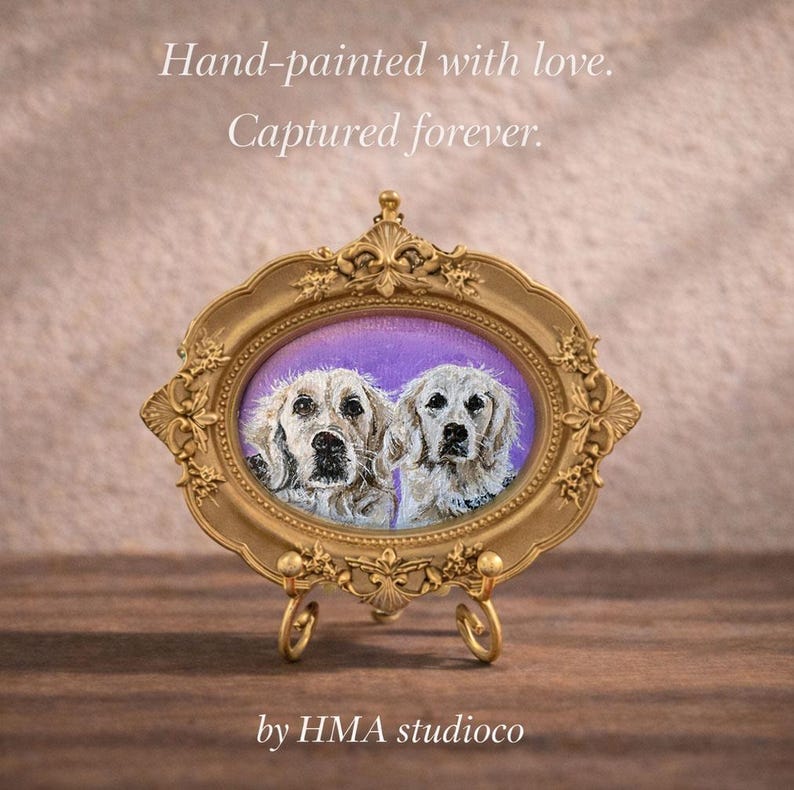 May include: An oval gold-toned ornate frame containing a painting of two light-colored dogs against a purple background. The frame rests on a small metal stand. Text above reads: Hand-painted with love. Captured forever. Text below reads: by HMA studioco.