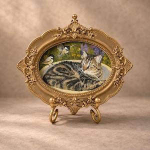 May include: An oval-shaped painting of a brown tabby cat resting in a bowl, accompanied by two small birds, set within an ornate, metallic gold-colored frame with intricate floral detailing, supported by a matching gold-toned metal display stand.