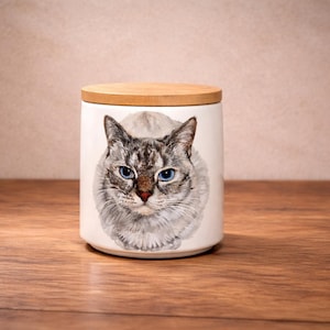 May include: A white ceramic cylindrical canister with a light brown wooden lid. The front of the canister features a detailed, realistic illustration of a cat with grey and white fur, pointed ears, and striking blue eyes, centered on the surface.