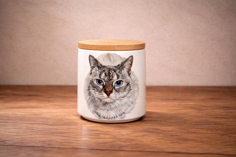 May include: A white ceramic cylindrical canister with a light brown wooden lid. The front of the canister features a detailed, realistic illustration of a cat with grey and white fur, pointed ears, and striking blue eyes, centered on the surface.