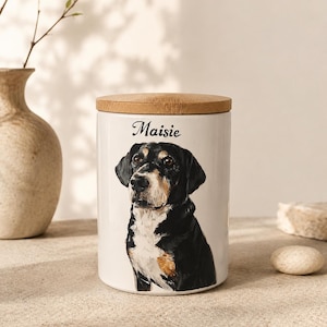 May include: A white ceramic cylindrical storage canister with a light brown wooden lid. The front of the canister features a painted illustration of a black, white, and tan dog with the name Maisie printed in black script lettering above the dog.