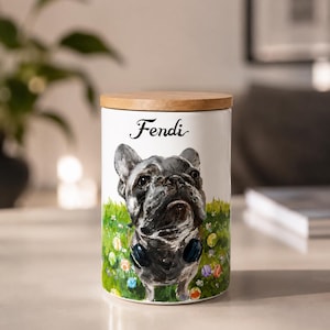 May include: A white ceramic cylindrical canister with a wooden lid, featuring a painted illustration of a grey French Bulldog wearing blue headphones, standing in a green grassy field with colorful balls, and the name Fendi written in black script.